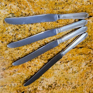 May include: Five stainless steel butter knives arranged diagonally on a speckled, golden-brown surface. The knives have a simple, elegant design with a slightly curved blade and a decorative handle.