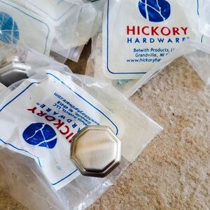 May include: Silver-toned octagonal cabinet knobs in clear plastic packaging. The packaging features the "Hickory Hardware" logo and company information. The knobs are on a light brown surface.