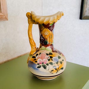May include: A ceramic pitcher with a floral design. The pitcher is decorated with a variety of colors, including yellow, blue, pink, green, and brown. The pitcher has a wide mouth and a handle that is shaped like a twisted rope.