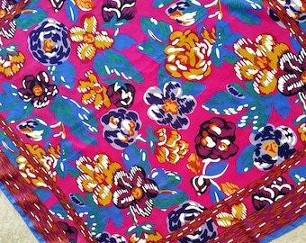 May include: A pink fabric with a floral pattern. The flowers are in shades of yellow, blue, and purple. The fabric has a border of brown and blue.