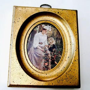 May include: A gold-colored oval picture frame with a raised edge, featuring a vintage image of a woman and a child in a garden setting.