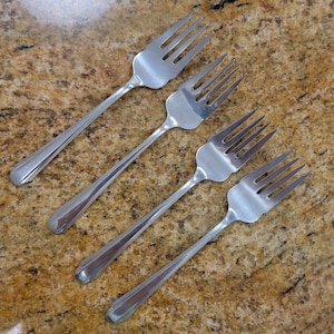 May include: Four stainless steel forks arranged diagonally on a speckled, brown surface. Each fork has a long, slender handle with a decorative design near the base and four tines. The forks are evenly spaced and angled.