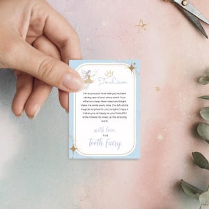 May include: A light blue Tooth Fairy card with gold accents and text. The card features a fairy illustration and the words "To Liam" and "The Tooth Fairy". The card is held by a hand.
