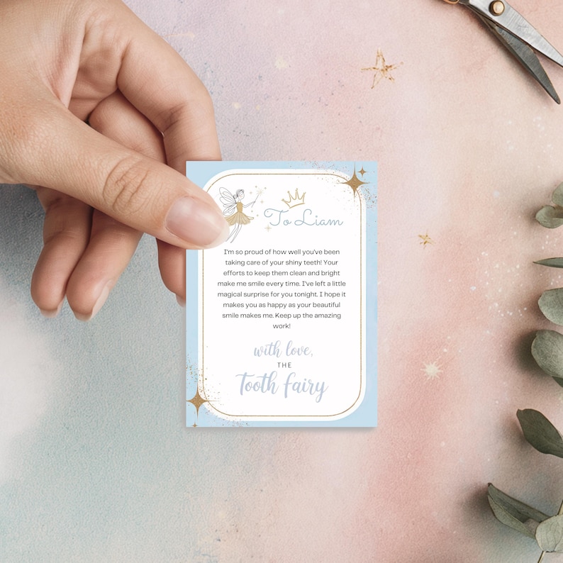 May include: A light blue Tooth Fairy card with gold accents and text. The card features a fairy illustration and the words "To Liam" and "The Tooth Fairy". The card is held by a hand.