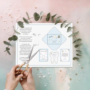 May include: A light blue Tooth Fairy kit with instructions, envelope, certificate, and receipt. The kit includes a diagram of how to fold the envelope. Gold scissors are cutting the paper. Eucalyptus and star decorations surround the kit.