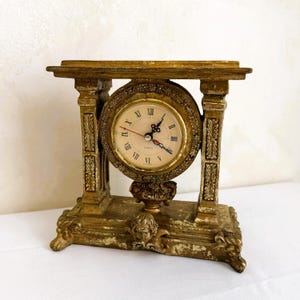 May include: A gold-colored, antique-style clock with Roman numerals on the clock face. The clock is supported by ornate pillars and a decorative base, creating a vintage aesthetic. The clock's hands indicate the time.