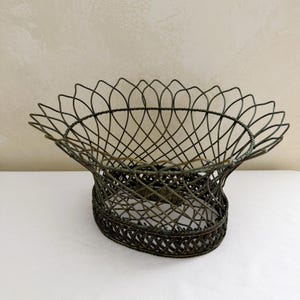 May include: An oval-shaped wire basket featuring a dark bronze finish. The structure consists of a woven base with a flared, open-work upper section that creates a scalloped rim. The metal wire is thin and curved, forming a lattice pattern throughout.