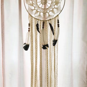 May include: A cream-colored dreamcatcher with a circular, crocheted web design. The dreamcatcher features hanging cream-colored ribbons, wooden beads, and white and black feathers. The circular frame is made of metal.