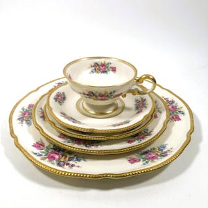 May include: A vintage tea set featuring a teacup, saucer, and plates. The set has a cream-colored base with floral patterns in pink, purple, and blue. Gold trim accents the edges of the cup and plates, adding a touch of elegance to the design.