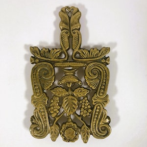 May include: Ornate brass trivet with a decorative floral and grape design. The trivet features a rectangular shape with a handle and intricate scrollwork. The brass has a warm, antique finish, adding to its vintage appeal. A functional and decorative kitchen accessory.