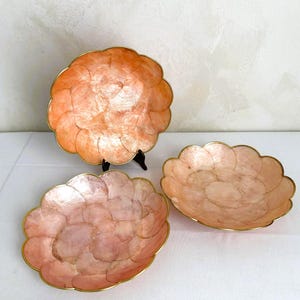 Puede incluir: Tres cuencos decorativos con forma de concha, bordes festoneados y bordes dorados. Los cuencos son en tonos melocotón y rosa. Un cuenco está sobre un soporte negro. Los cuencos son probablemente para fines decorativos.