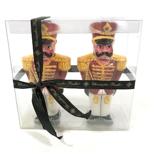 May include: Two ceramic nutcracker figurines in a clear plastic box, tied with a black ribbon. The nutcrackers are dressed in red and gold uniforms with black mustaches and hats. The ribbon has the text "Christopher Radko".