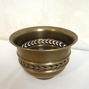 May include: A vintage brass bowl with a decorative leaf pattern around the middle and a perforated design near the top. The bowl has a wide, rounded rim and a slightly flared base. The metal has a warm, aged patina.