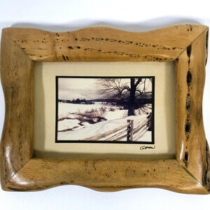 May include: A framed landscape photograph featuring a winter scene with snow-covered fields, a wooden fence, and a bare tree. The frame is made of light-colored wood with a unique, wavy edge.