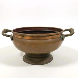 May include: A copper bowl with two ornate handles and a pedestal base. The bowl has a wide, open top and a reddish-brown interior. The handles are detailed with a decorative design. The bowl is likely vintage or antique.