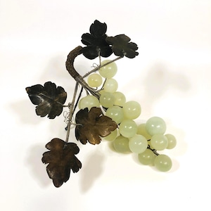 May include: A decorative sculpture of a bunch of grapes. The grapes are a pale green color and appear to be made of a smooth material. The leaves are dark brown and the stem is a lighter brown. The sculpture is set against a white background.