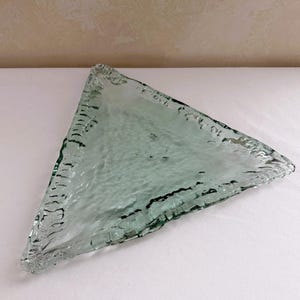 May include: A triangular glass dish with a pale green tint and a textured, rippled surface. The edges of the dish are thick and uneven, featuring a rough, jagged appearance that contrasts with the smoother, wavy center of the glass piece.