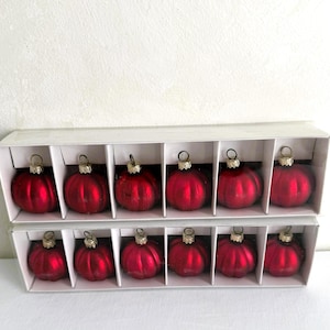 May include: A set of twelve red, ribbed, spherical glass ornaments arranged in two rows within a white cardboard box. Each ornament features a metallic gold-toned cap with a small loop for hanging, showcasing a consistent glossy finish.