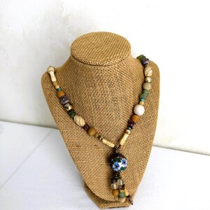 May include: A handcrafted necklace featuring a variety of beads in shades of beige, brown, green, and blue. The necklace includes wooden beads, stone beads, and a central pendant with a floral design. The necklace is displayed on a burlap bust.