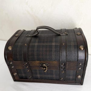 May include: A small, vintage-style chest with a plaid pattern on the top and sides. The chest is made of wood and leather, with metal studs and a handle. The clasp is a decorative metal piece. The overall color scheme is dark brown and black.