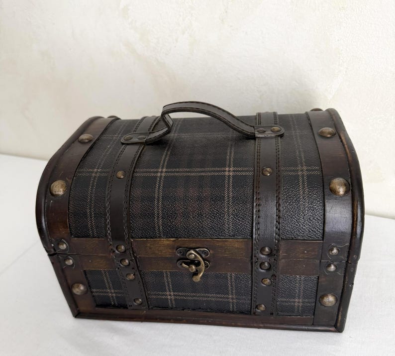 May include: A small, vintage-style chest with a plaid pattern on the top and sides. The chest is made of wood and leather, with metal studs and a handle. The clasp is a decorative metal piece. The overall color scheme is dark brown and black.