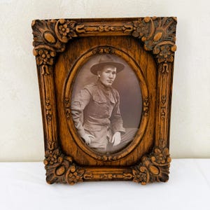 May include: A rectangular brown wooden picture frame with ornate floral carvings at the corners and an oval center opening. Inside the frame is a sepia-toned photograph of a person wearing a military uniform and a wide-brimmed hat, seated in profile.