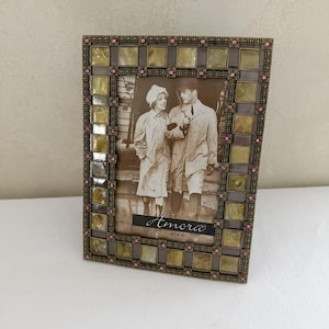 May include: A rectangular picture frame with a metallic border featuring square, iridescent yellow-green tiles and small pink rhinestones. The frame holds a black and white photograph of two people with the text Amora 4x6 printed at the bottom.