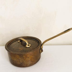 以下が含まれることがあります： 蓋と長い柄が付いた、小さなアンティークの銅製ソースパン。鍋は変色した古びた外観で、真鍮製のハンドルと蓋のハンドルが付いています。ハンドルは鍋の側面にリベットで留められています。