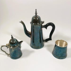 May include: A silver-plated tea set with a teapot, creamer, and sugar bowl. The teapot has a black handle and a domed lid. The creamer and sugar bowl have a silver-plated finish.