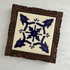 May include: A square ceramic tile featuring a dark blue symmetrical floral pattern on an off-white background, encased in a dark brown wooden frame with carved decorative details along the edges and corners of the square border.