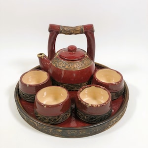 May include: A red ceramic tea set with a teapot and four cups, all resting on a matching tray. The teapot has a decorative handle and lid, with gold-colored accents. The cups and tray have a dark brown decorative border.