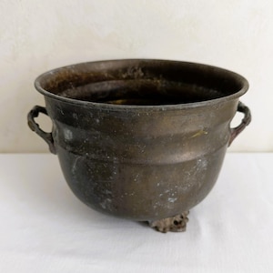 May include: A round, dark bronze-toned metal pot featuring a wide, open top, two small decorative side handles, and three small feet at the base. The surface of the metal shows a weathered, aged patina with visible variations in color and texture.