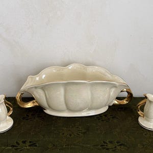 Pode incluir: Uma travessa de cerâmica creme com alças e acabamentos dourados, ladeada por dois pequenos jarros a condizer. O conjunto tem uma borda ondulada e assenta numa superfície verde escuro.