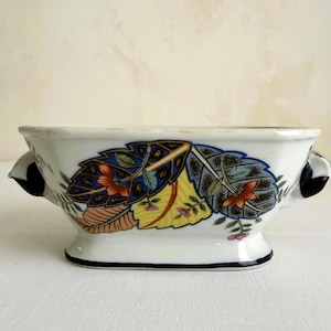 May include: A white ceramic bowl with a low profile and two black handles. The bowl is decorated with colorful leaf and floral patterns in blue, yellow, orange, and pink. The base and rim are outlined in dark blue.