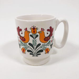 May include: White ceramic mug with a folk art design featuring two orange birds with red and green accents. The birds are perched on a stylized green plant with red flowers.