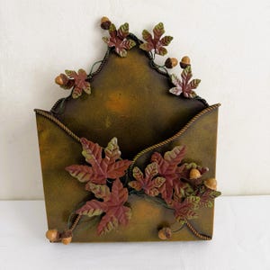 May include: A wall-mounted metal mail holder shaped like an envelope with a dark olive green and bronze finish. The piece is decorated with three-dimensional metal maple leaves in shades of red and green, along with small brown acorn accents.