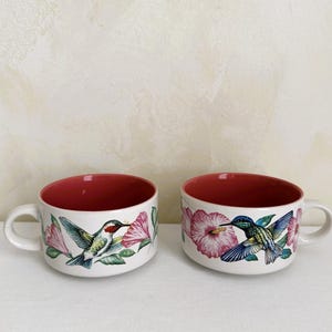 May include: Two white ceramic mugs with red interiors. Each mug features a colorful illustration of a hummingbird and pink flowers. The mugs have a curved shape and a handle.
