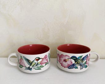 May include: Two white ceramic mugs with red interiors. Each mug features a colorful illustration of a hummingbird and pink flowers. The mugs have a curved shape and a handle.