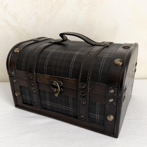 May include: A small, dark brown chest with a plaid pattern on the front and top. The chest has a handle, metal studs, and a clasp closure. The chest is made of wood and leather.