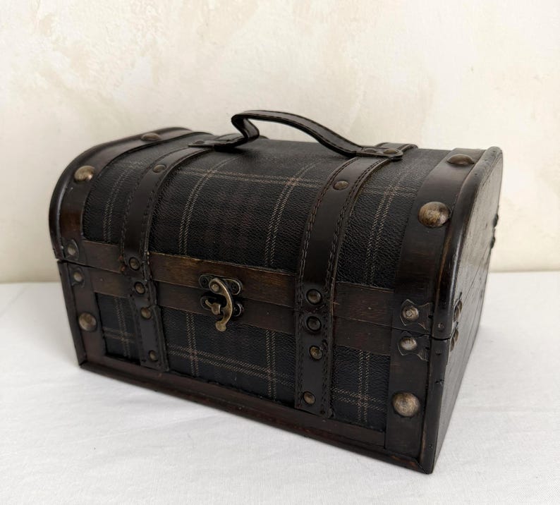 May include: A small, dark brown chest with a plaid pattern on the front and top. The chest has a handle, metal studs, and a clasp closure. The chest is made of wood and leather.