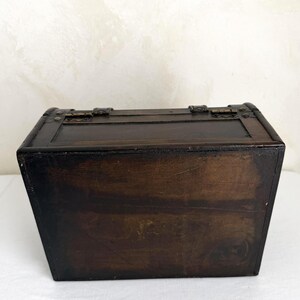 May include: A small, dark brown wooden chest with metal hardware. The chest has a rectangular shape with a hinged lid and metal clasps. The wood grain is visible, and the chest appears to be aged.