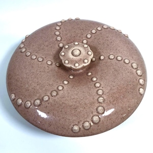 Pode incluir: Tampa redonda de cerâmica salpicada de cor malva, com um botão floral central. A tampa é decorada com linhas pontilhadas em relevo que irradiam do botão. O botão e os pontos são de uma cor creme mais clara.