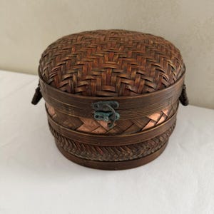 May include: A round, lidded storage container crafted from dark brown woven bamboo strips. The container features a metal latch closure on the front and small woven handles on the sides. The exterior displays a multi-layered, intricate herringbone weave pattern with visible copper-colored accents between the woven sections.