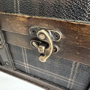 May include: Close-up of a decorative chest with a plaid pattern. The chest features a dark brown wooden frame and a metal clasp. The clasp is a bronze color and is secured with small screws. The chest's exterior is black with a white plaid pattern.