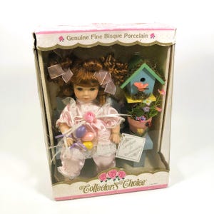 Puede incluir: Muñeca de porcelana bisque Collector's Choice en una caja de presentación. La muñeca tiene cabello castaño rizado, ropa rosa y sostiene huevos de Pascua. También hay una pajarera en miniatura y flores en la caja. La caja dice "Genuine Fine Bisque Porcelain".