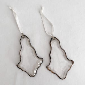 May include: Two clear, irregular, organic-shaped glass ornaments with smooth, translucent surfaces. Each piece features a dark, metallic-toned soldered edge and is suspended by a thin, white satin ribbon loop attached to the top of the glass.