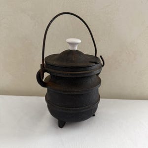 May include: A dark brown cast iron pot with a tiered, rounded body, three small feet, and a matching lid featuring a white ceramic knob. A thin, arched metal handle is attached to the sides, and the lid includes a small notch for a utensil.