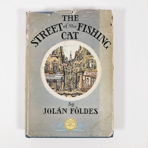 Puede incluir: La portada de un libro azul titulado "The Street of the Fishing Cat" de Jolán Földes. La portada presenta una ilustración de una escena callejera en una ciudad europea con edificios, tiendas y un gato.