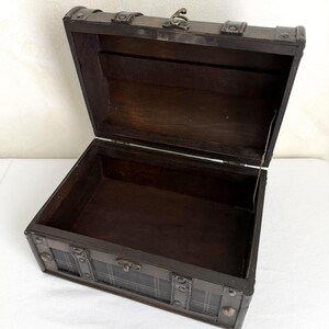 May include: An open, dark brown wooden chest with metal accents. The chest has a hinged lid and a plaid pattern on the front. The interior is empty and the chest is sitting on a white surface.