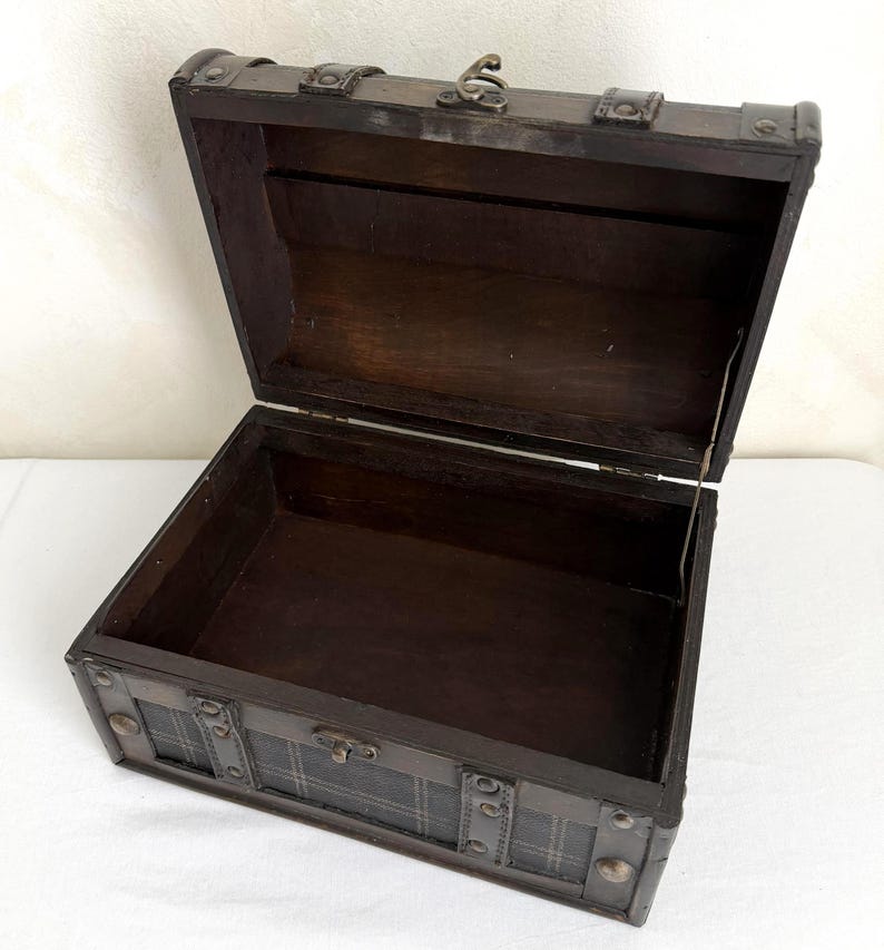May include: An open, dark brown wooden chest with metal accents. The chest has a hinged lid and a plaid pattern on the front. The interior is empty and the chest is sitting on a white surface.